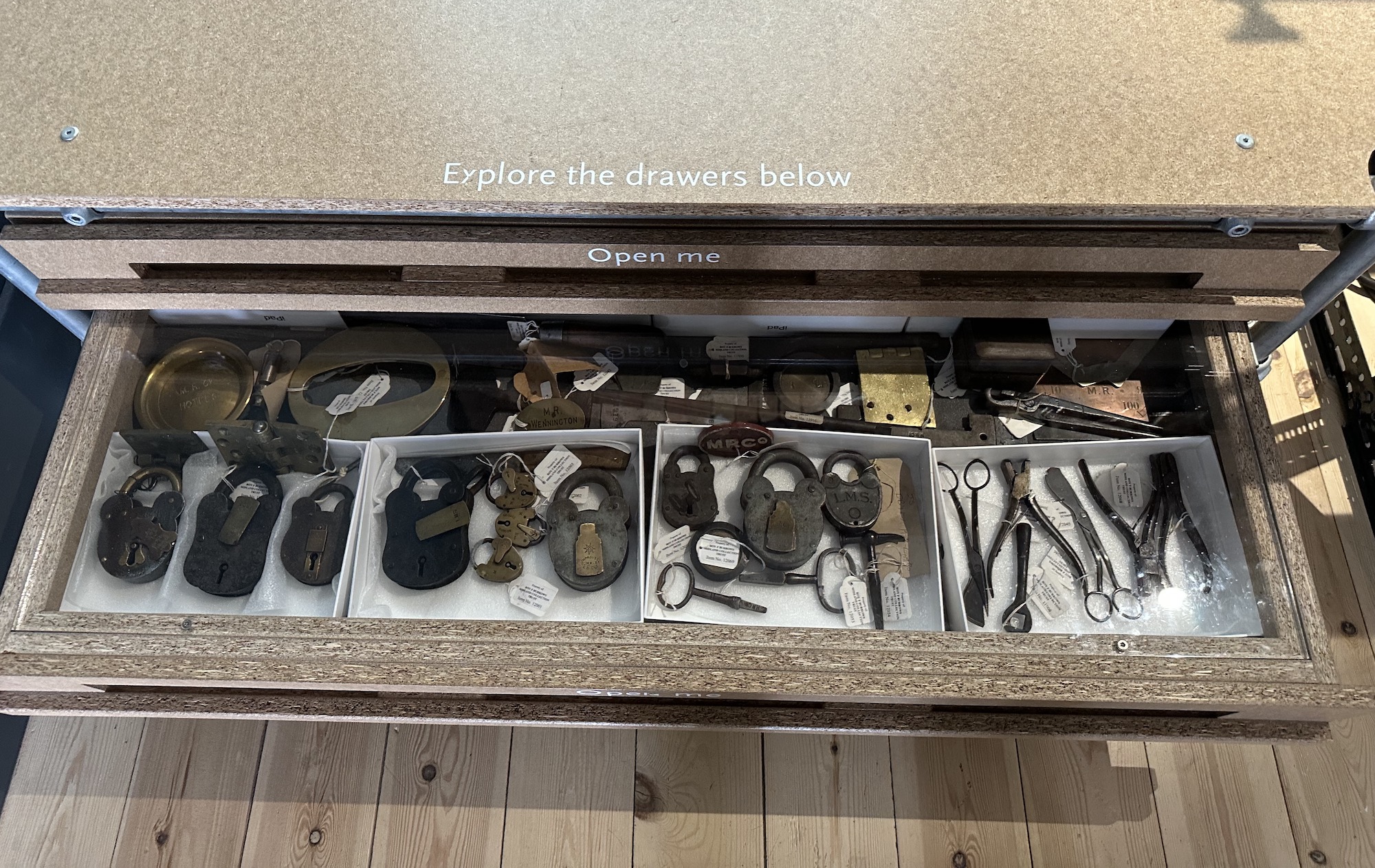Looking down into a perspex covered drawer on the Assemblage of the Museum of Making. A large collection of small items associated with the Midland Railway is in view; including padlocks, office equipment, ticket clippers, a carriage door key, brass hinges... all stamped M.R. Co. to make its ownership clear. These are mostly now contained in white boxes to prevent them moving about when the drawer is closed.