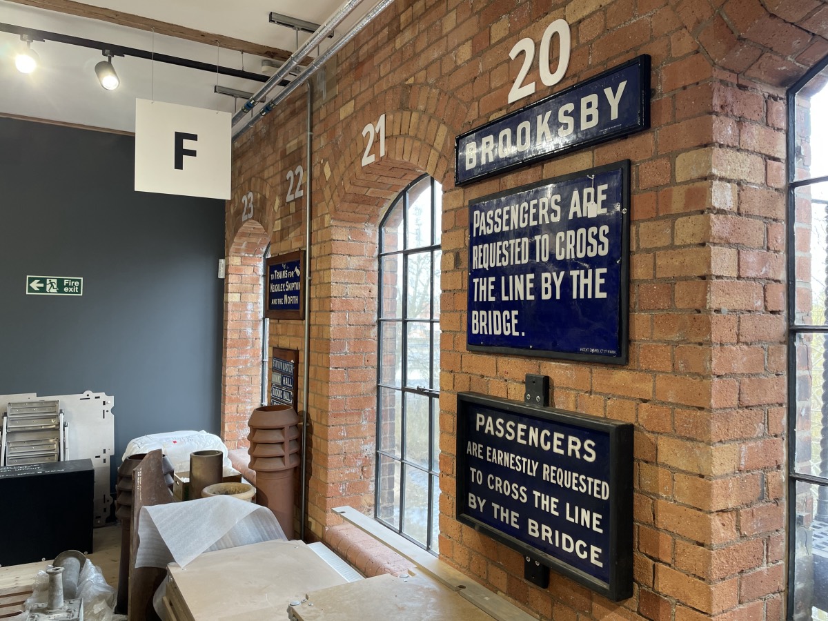 Blue enamel signs fixed to a bare brick wall; 'Brooksby' and two signs which each read 'Passengers are earnestly requested to cross the line by the bridge' 