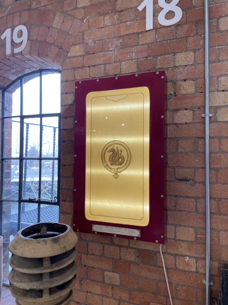 An opaque glass pane bearing the Midland Railway's wyvern device, mounted within a lightbox which is itself mounted on the bare brick wall of The Assemblage
