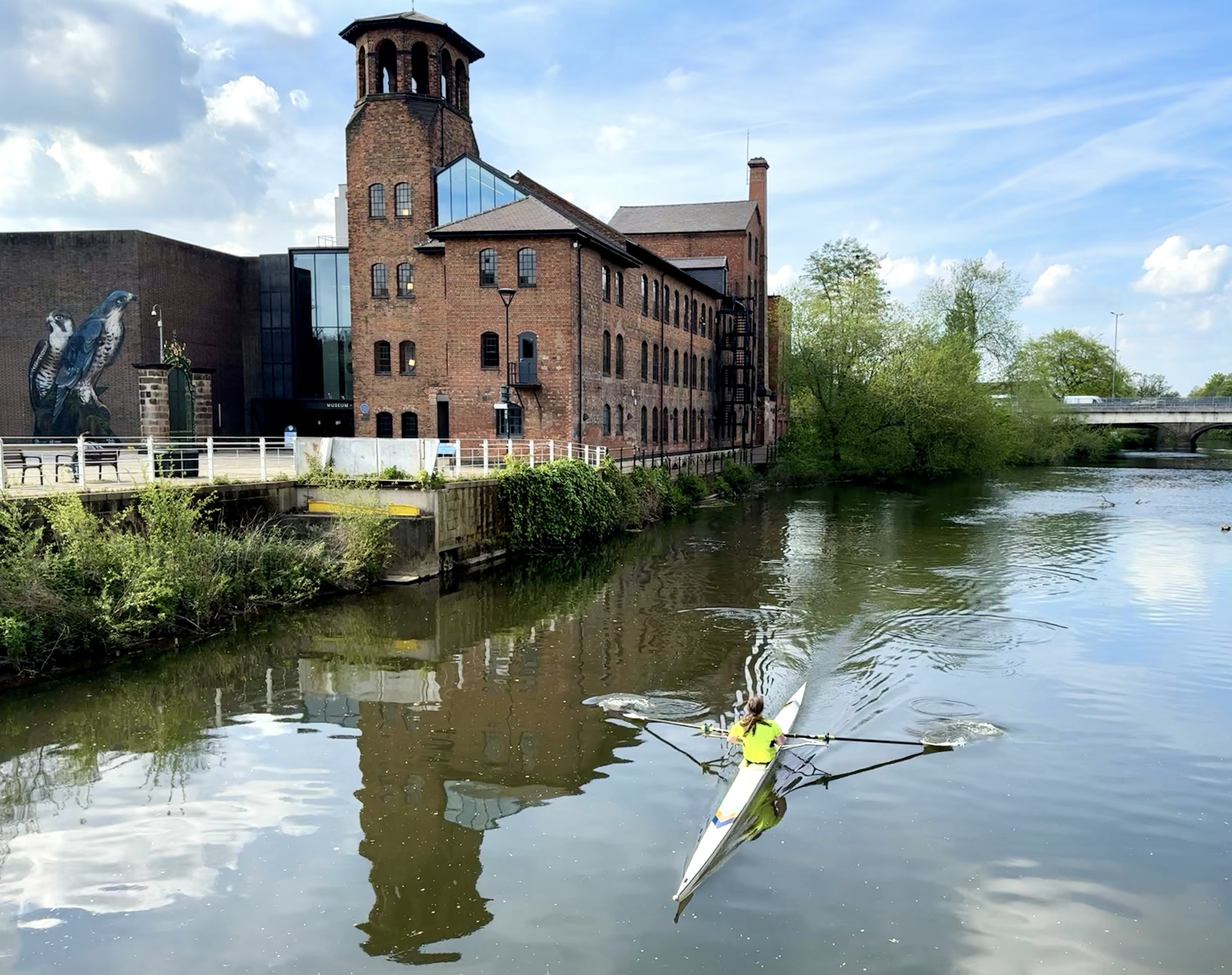 A view of the Silk Mill from the footbridge over the River Derwent with a single rower in the foreground.