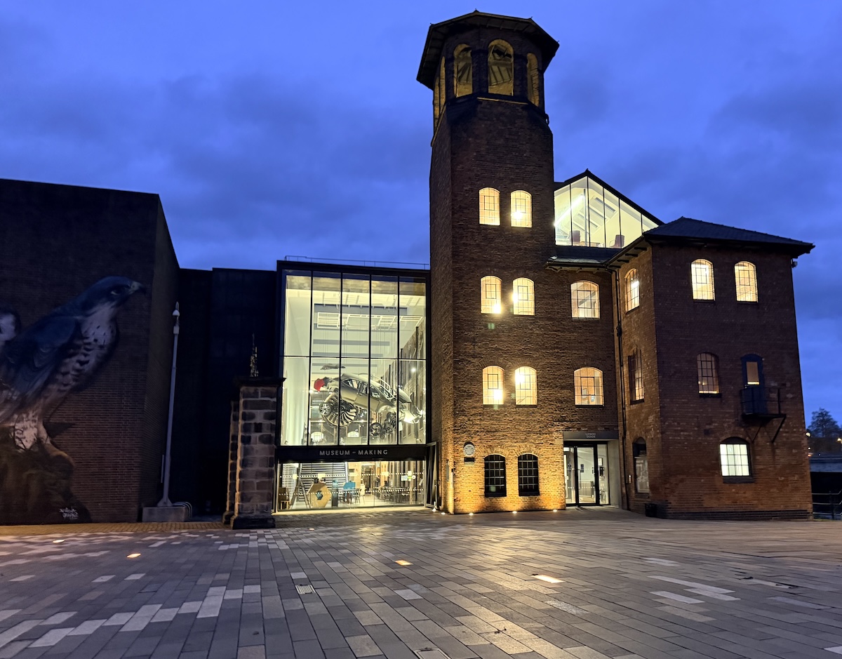 A winter's evening view of the Silk Mill at Derby, lit from inside and the lights of Cathedral Green. Alt: Brick building with lit tower and glass entrance at dusk. Large peregrine mural on adjacent wall. Open plaza in front. 
