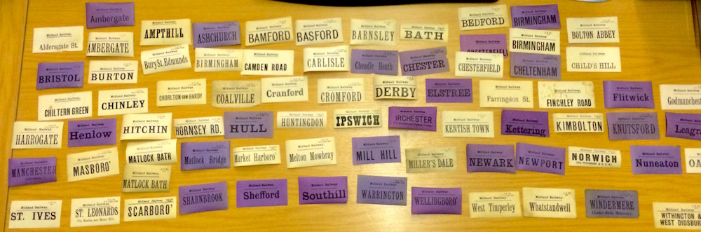 A collection of about two dozen rectangular luggage labels with the names of a wide variety of station names. Many are coloured purple, others are cream.