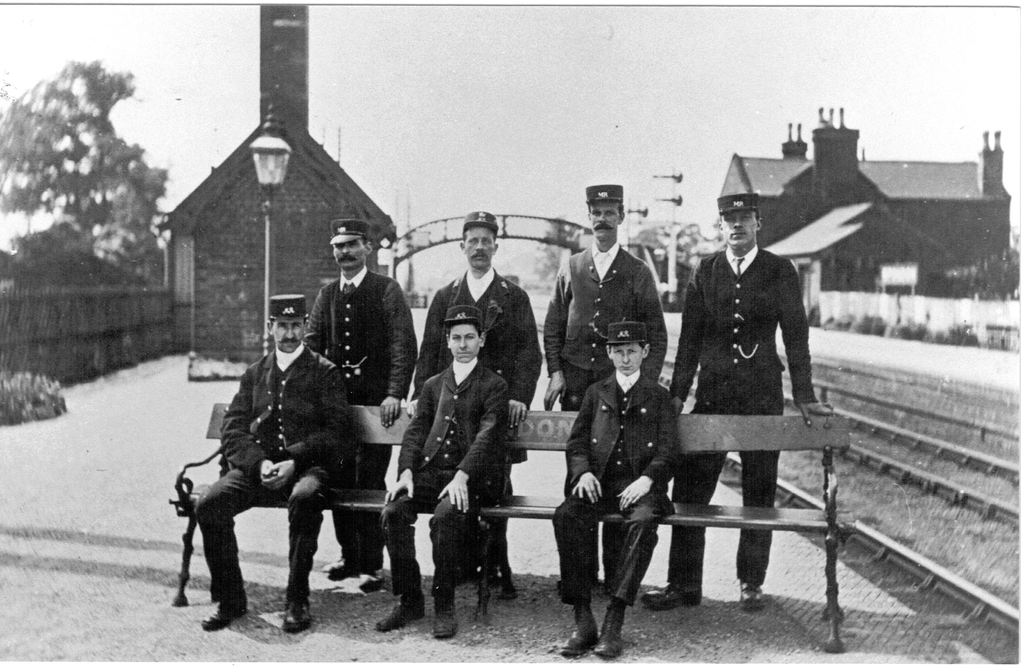 A photograph of seven uniformed Midland Railway employees ranging from barely teenaged to middle aged, three are sitting on one of these benches with the name Spondon just visible beteween them, the other four are standing behind