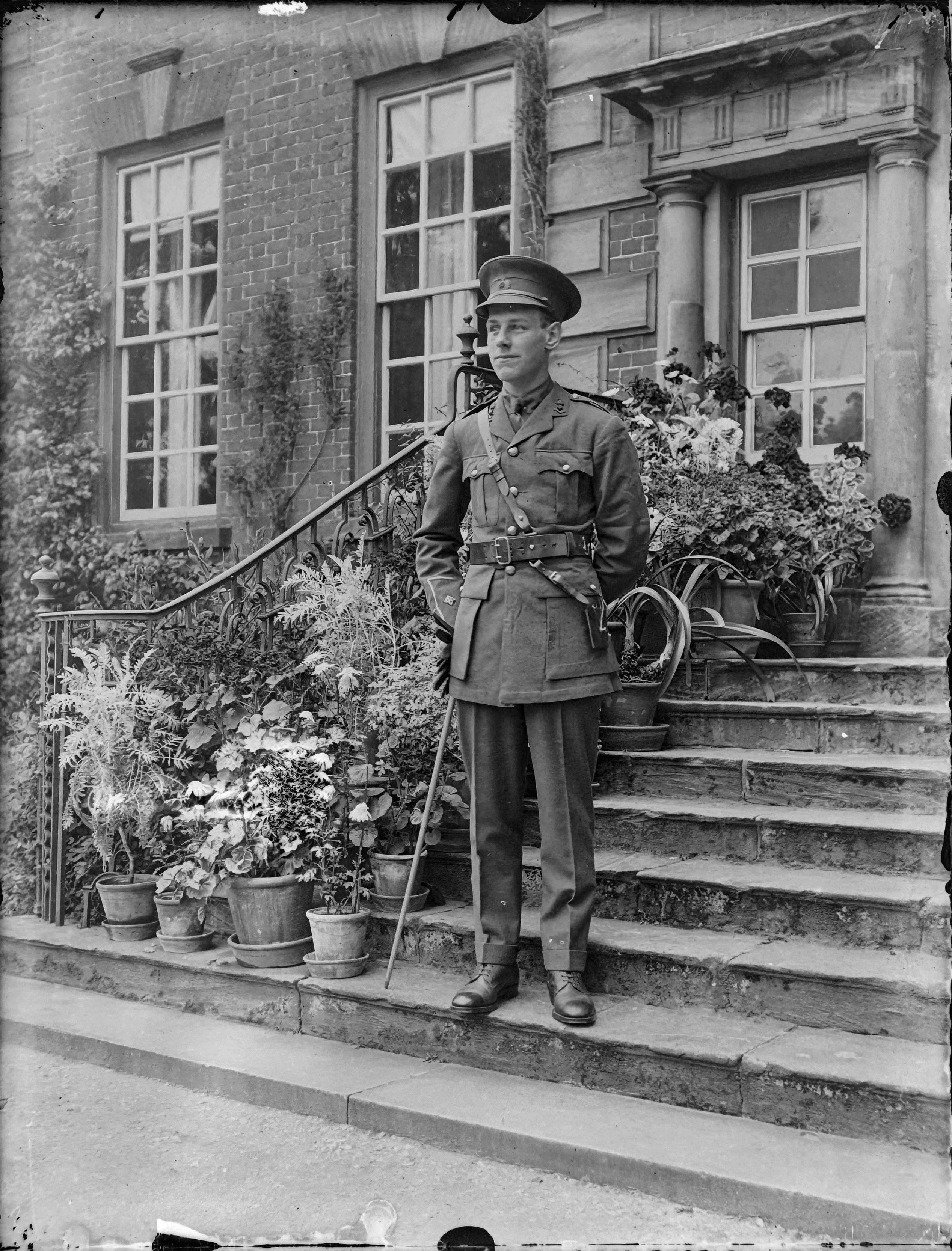 A young man in military uniform stands on the steps of a large house with many potted plants on the steps behind him