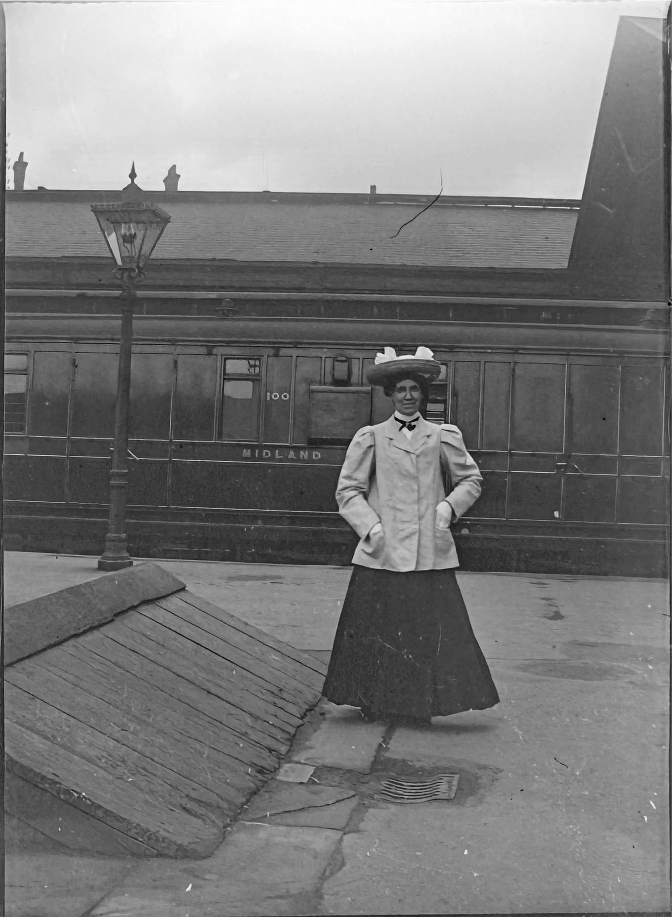 An Edwardian lady with a fancy hat, light jacket and long skirt stands on Derby Station posing for the camera with a railway carriage bearing the number 100 and Midland is seen behind her