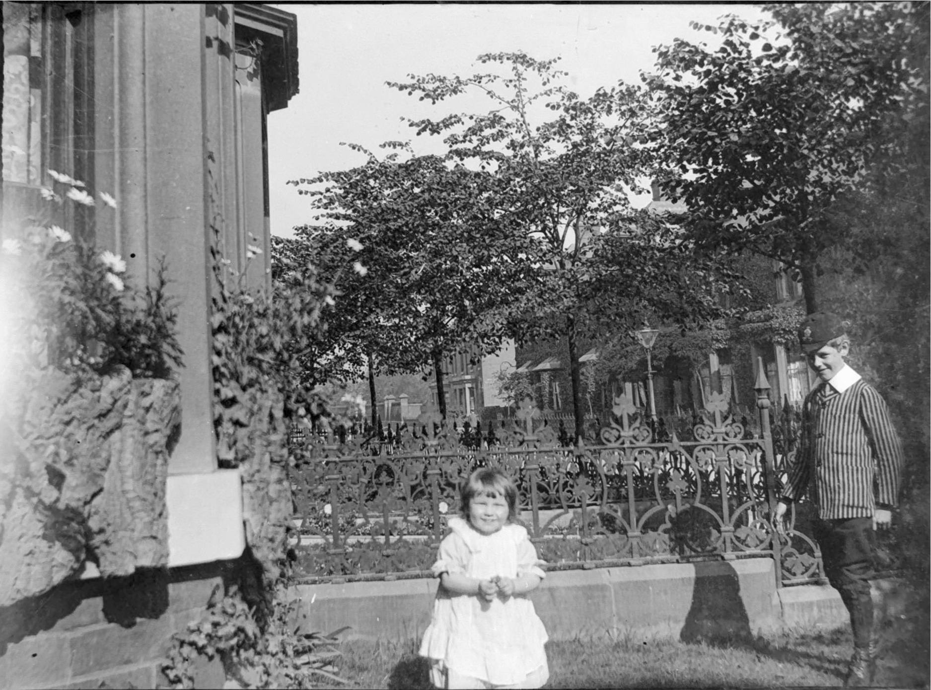 A very young girl and a slightly older boy in his smart school uniform play in the front garden of a house with a view of the street behind them