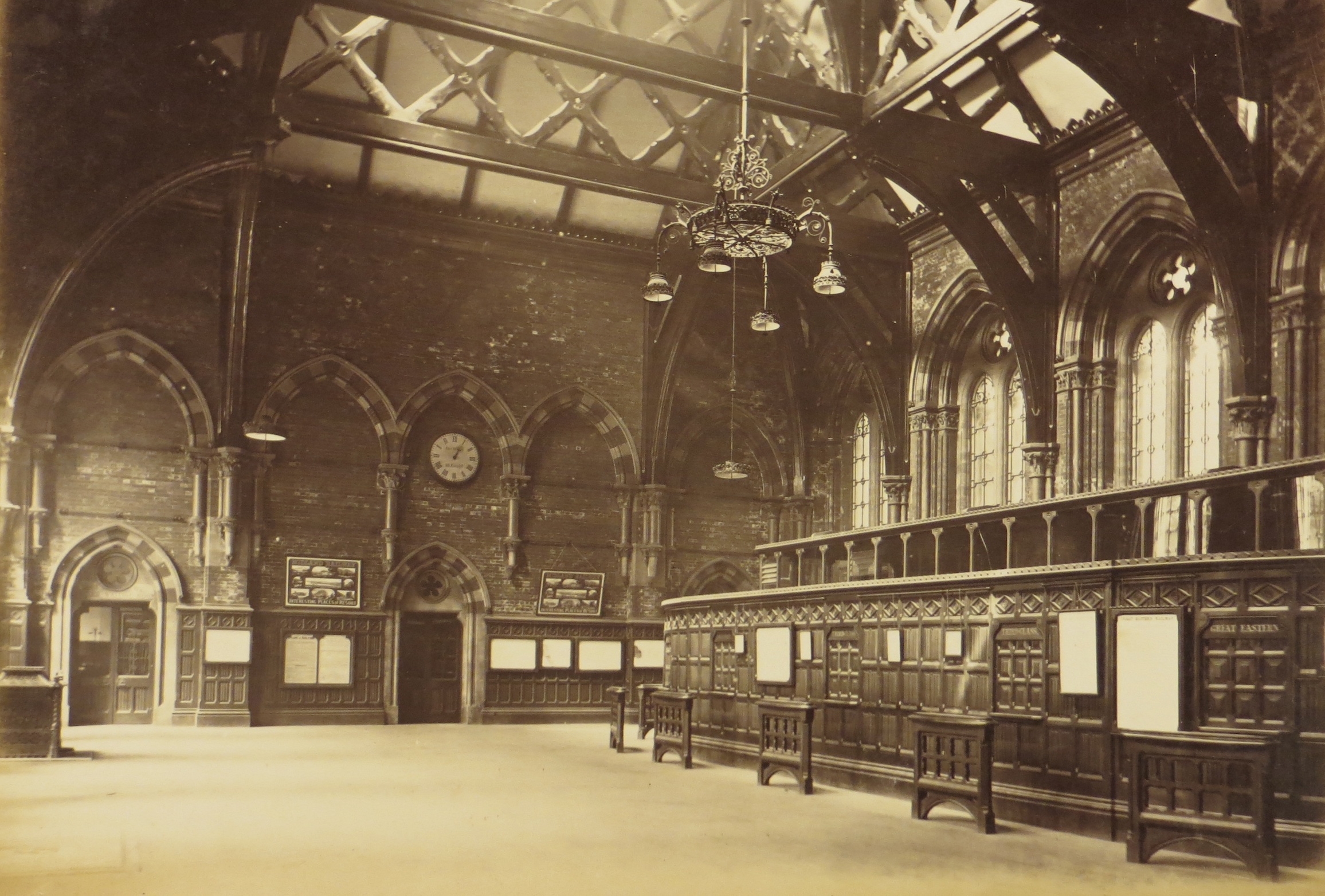 Black & white image: a cavernous and grand wood panelled but deserted booking hall with a series of closed ticket windows with their privacy barriers in front
