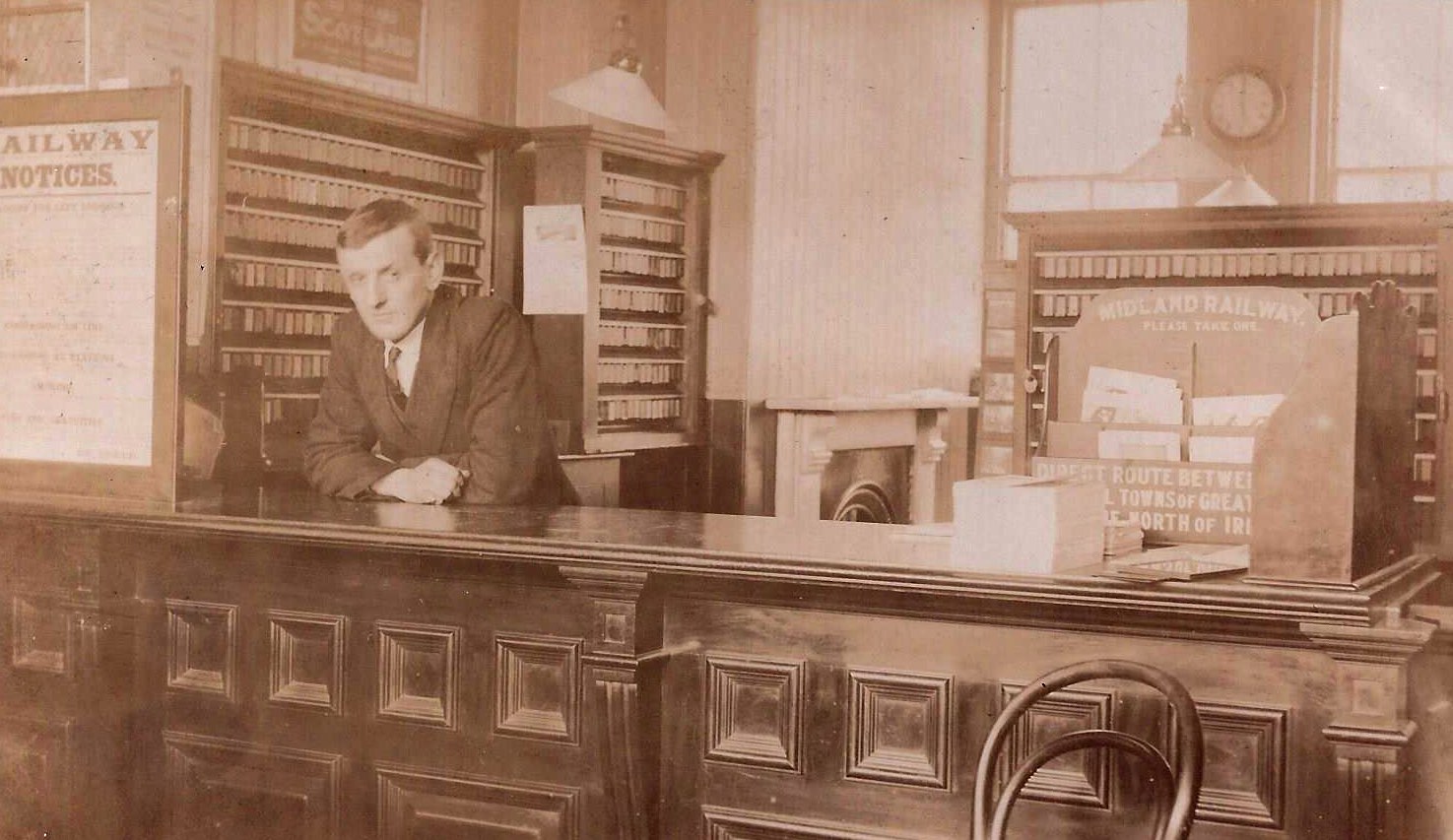 Black & white image: A rather stern looking clerk stares at the camera while leaning on the counter of a Midland Railway ticket office