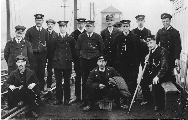 A group of 14 men all wearing Midland Railway uniforms pose for the camera. Beside them is a grounded coach body and a Midland Railway signal box is in the mid-distance behind.