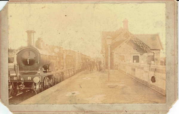 Johnson 4-4-0 tender engine stands at an unidentifed station with the staff watching the photographer from a distance. The station building is on the right and is notable for its ornate bargeboards.