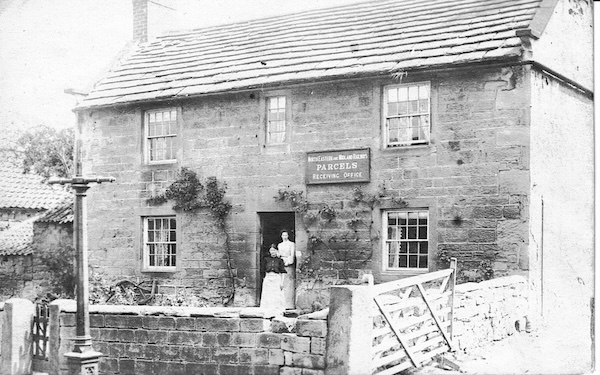 A stone built house with a lady and probably her daughter standing in the doorway posing for the photogrpher. A sign over the door reads North Eastern and Midland Railways Parcels Receiving Office