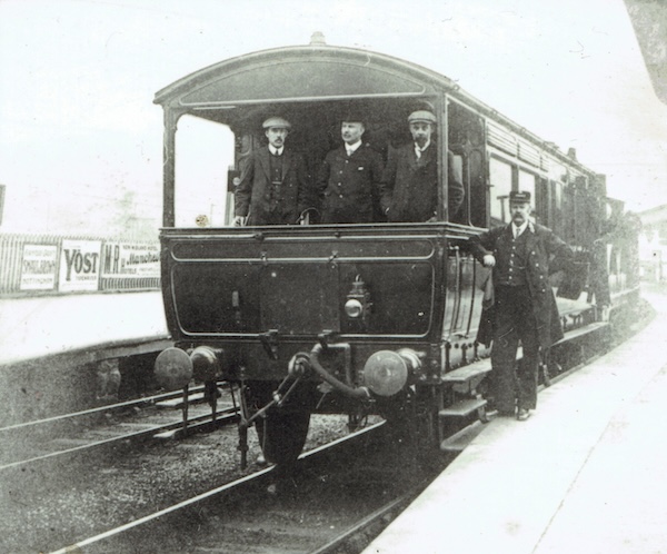 A Midland Railway six wheel Inspection Saloon stands in the platform at an unidentified station platform with two men in working class dress flanking a man in notably smarter attire. The Guard stands on the platform posing alongside the train.