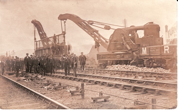 Postcard print of a photograph showing a Midland Railway permanent way gang posing for a photographer whilst two cranes are in the middle of lifting into place a cast iron bridge girder and decorative balustrade. The view includes three plank wagon No. 22556.