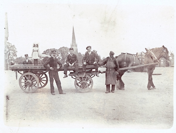 Photograph showing a group of Midland Railway staff, a young girl and what looks like a blacksmith posing in front of a Midland Railway dray and its horse. The location is unknown but is evidently a wide station yard with a distinctive church spire in the background.