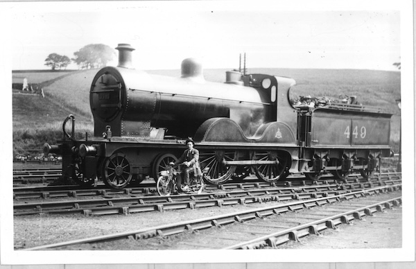 Midland Railway Compund locomotive No.449 is posed in an unidentified rural yard, possibly a running shed, with a man in civilian clothing astride a motorcycle in the ‘six foot’ adjacent to the front of the engine.