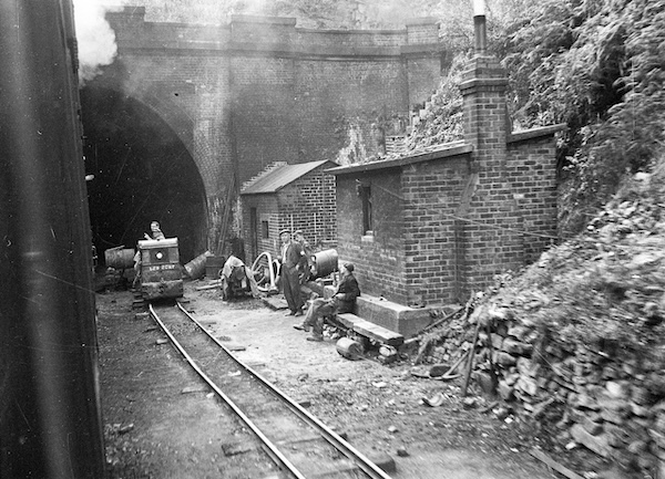 A group of workmen have paused while a passeneger train, from which the photograph has been taken, enters the tunnel they are working in. The work is extensive as there is a temporary narrow-gauge railway laid with one of the workmen is on a small petrol driven locomotive.