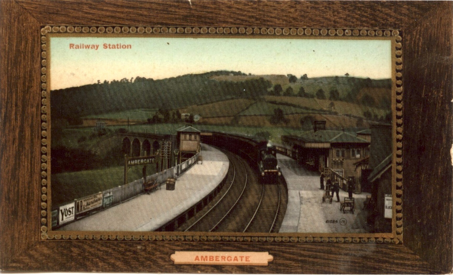 A colourised post card view of Ambergate station taken from the footbridge looking north west showing the two Manchester line platforms. On the left, three enamel adverting signs can be seen which exactly match our mystery photo
