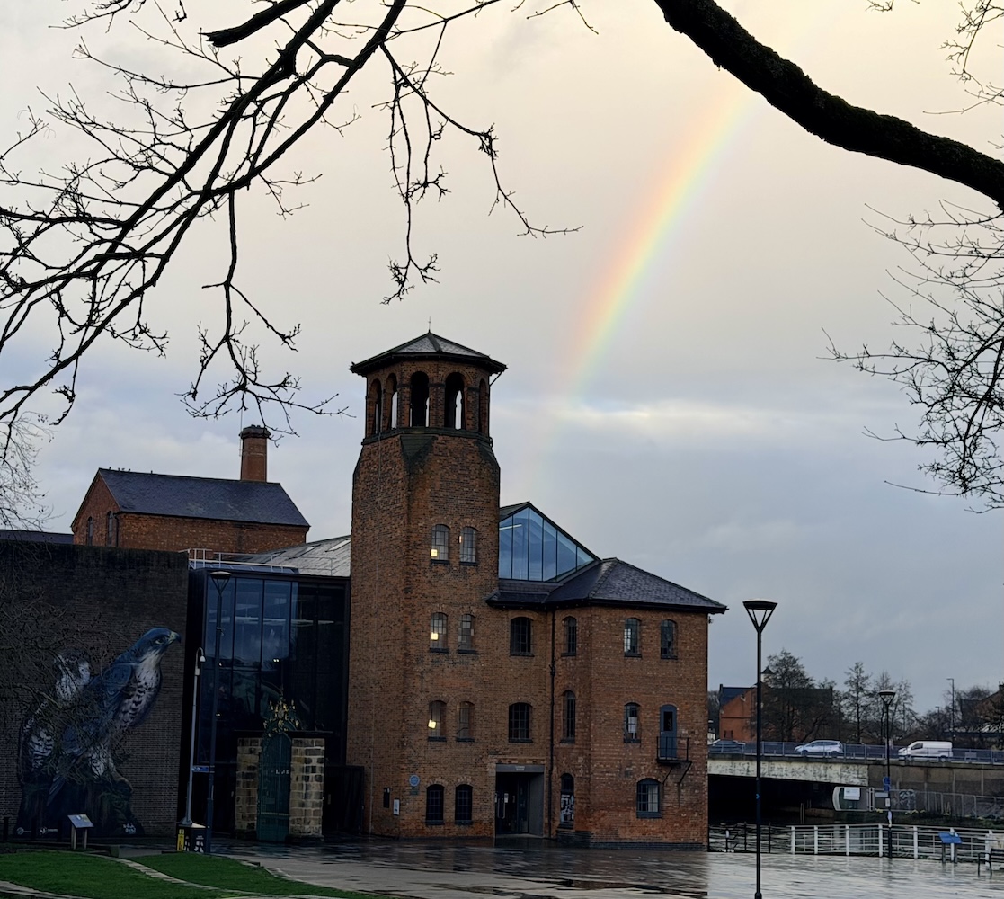 A brick tower with arched windows under a rainbow, next to a modern building with a falcon mural, on a wet day.