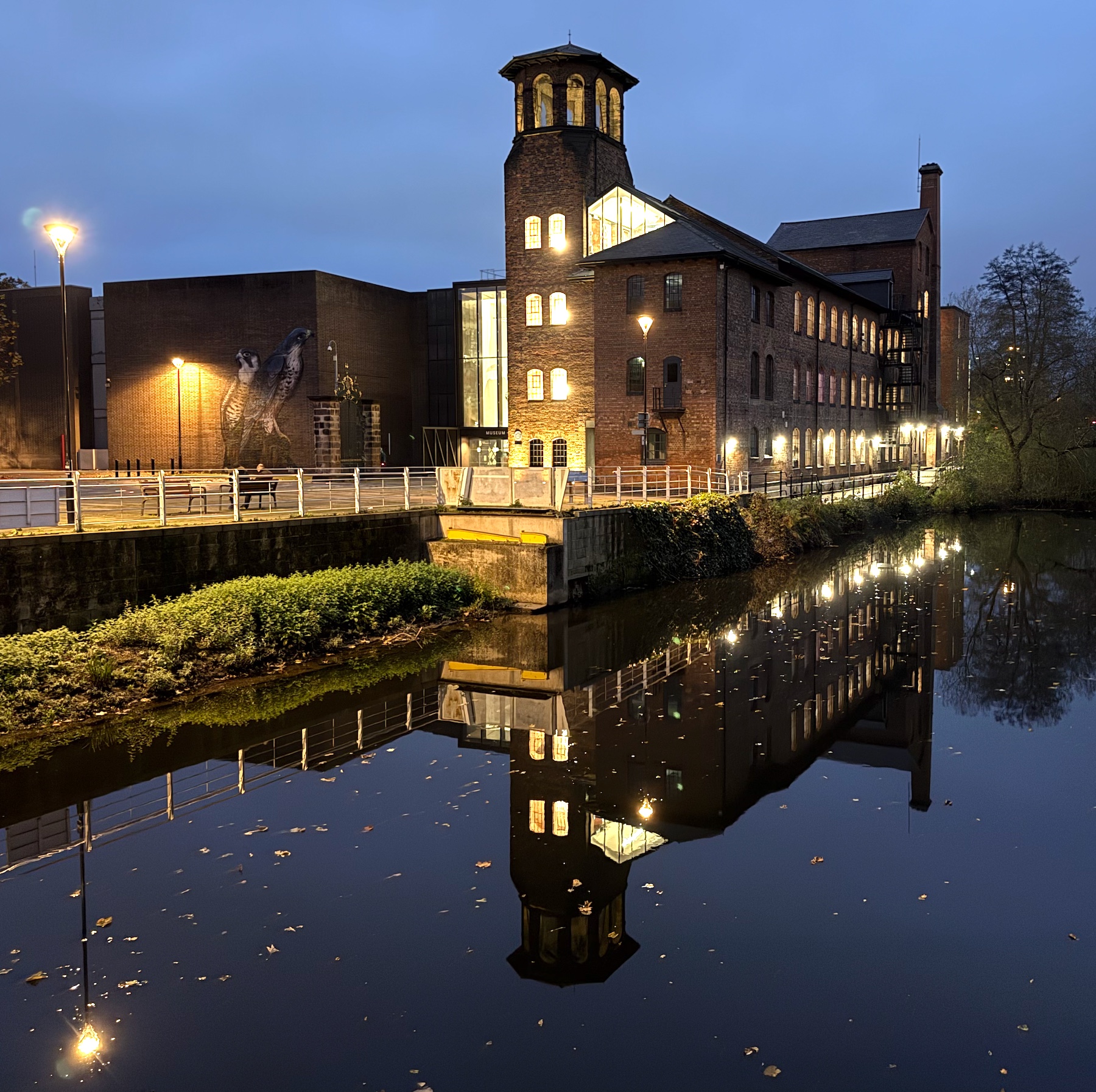 The Silk Mill all lit up in the late dusk viewed from the footbridge over the River Derwent which reflects the image in the water.