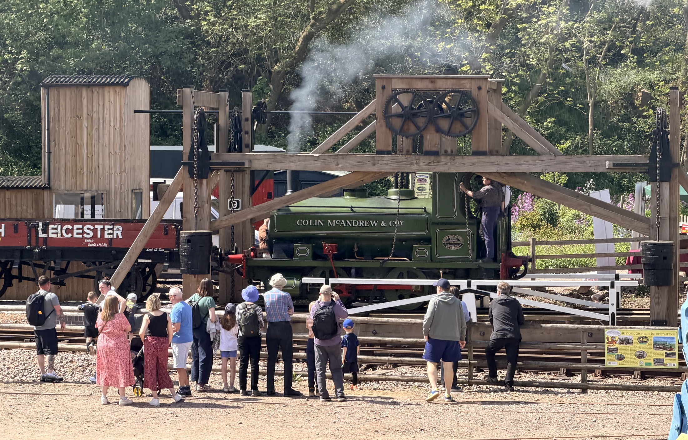 Green steam train with people watching behind a wooden fence; trees and a wooden building in the background.
