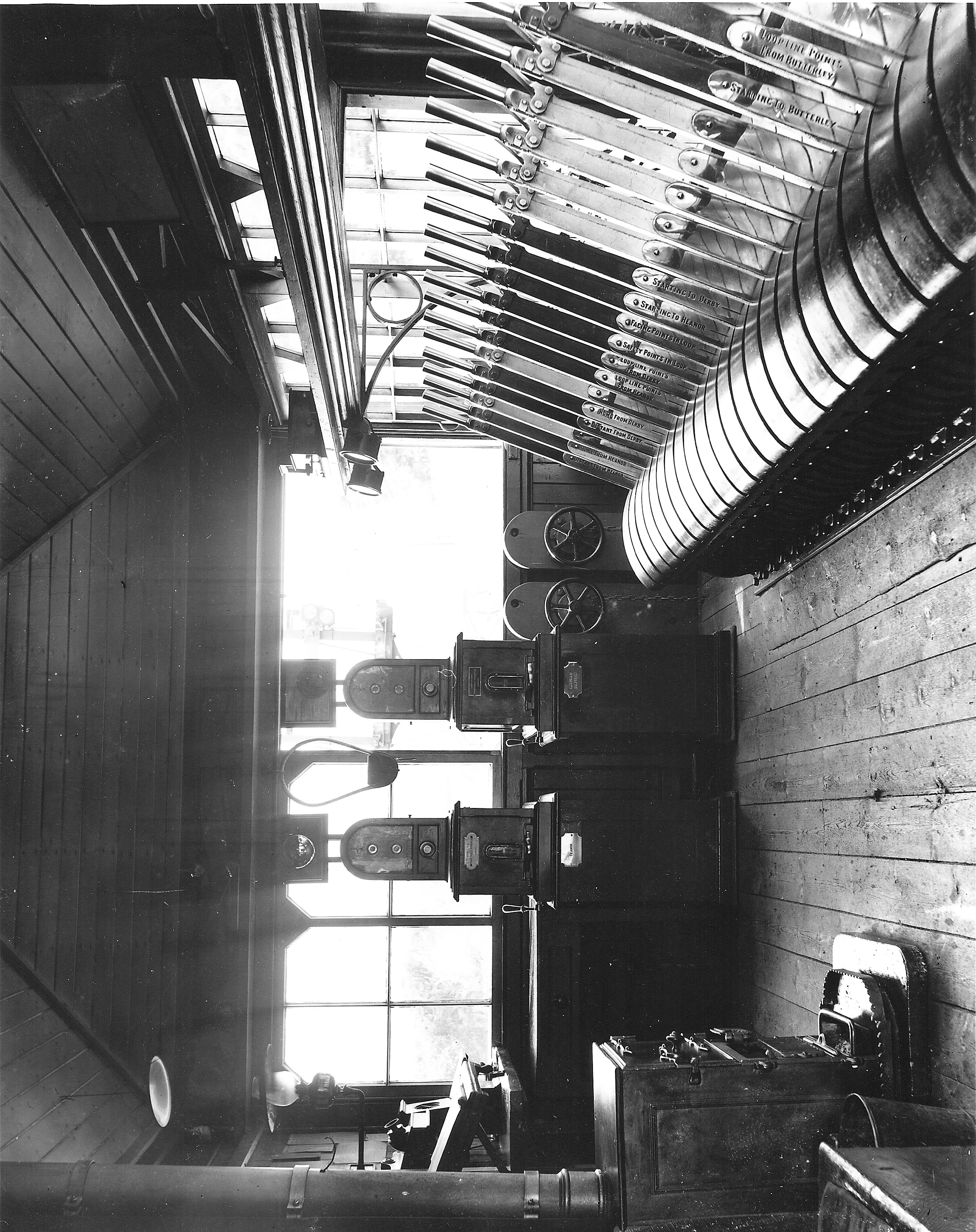 A photograph of the rather sparten interior of a signal box. The lever frame is to the right and a stove at the back of the box is to the left. We are looking at the end windows with a pair of large Tyers' tablet instruments on plinths dominate the view. The one on left bears a brass plaque that says Heanor while the other reads Marehay Junction.