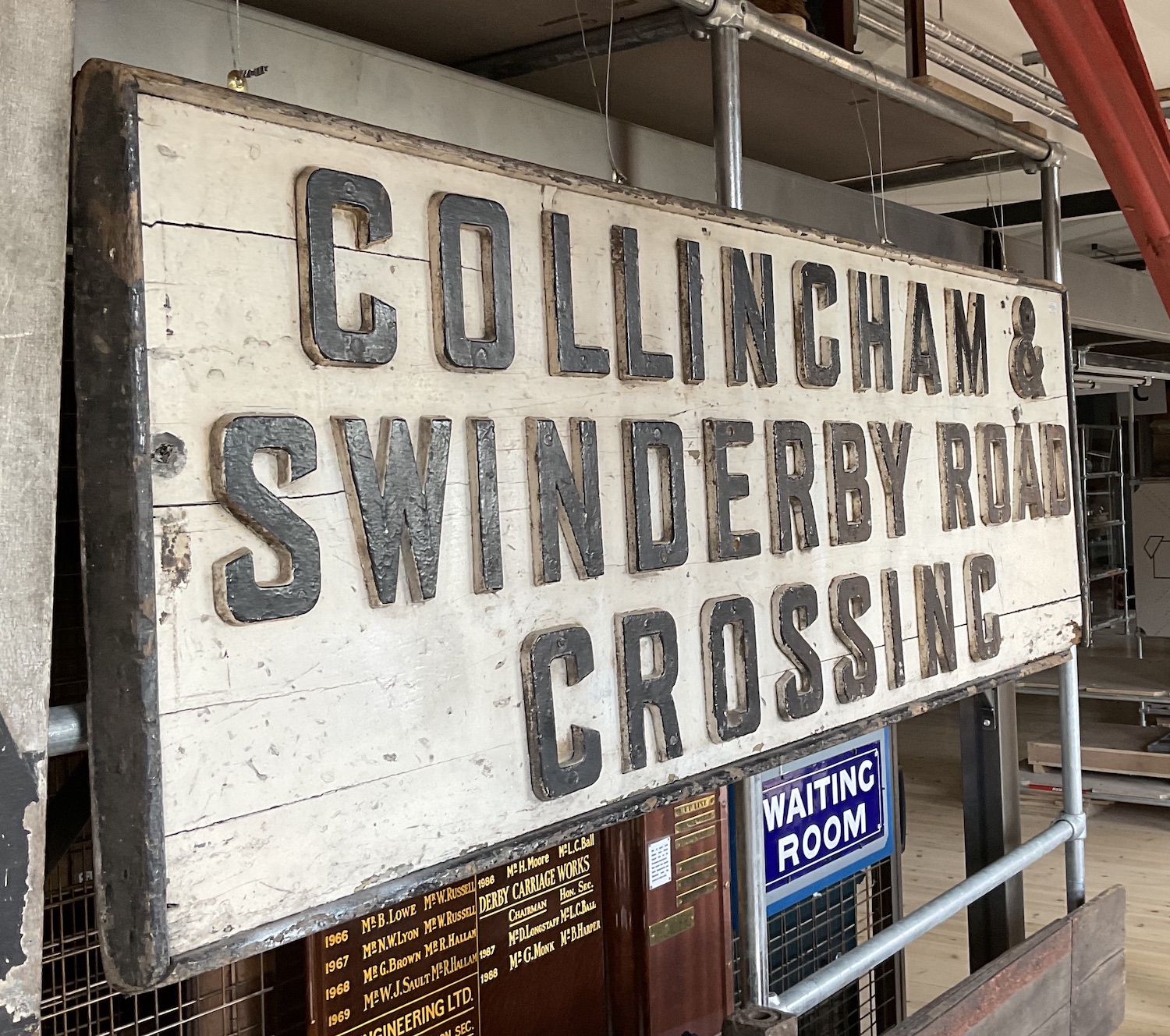 A large wooden board with beading around the edge, painted white with black letters spread over three lines which reads 'Collingham & Swinderby Road Crossing' 